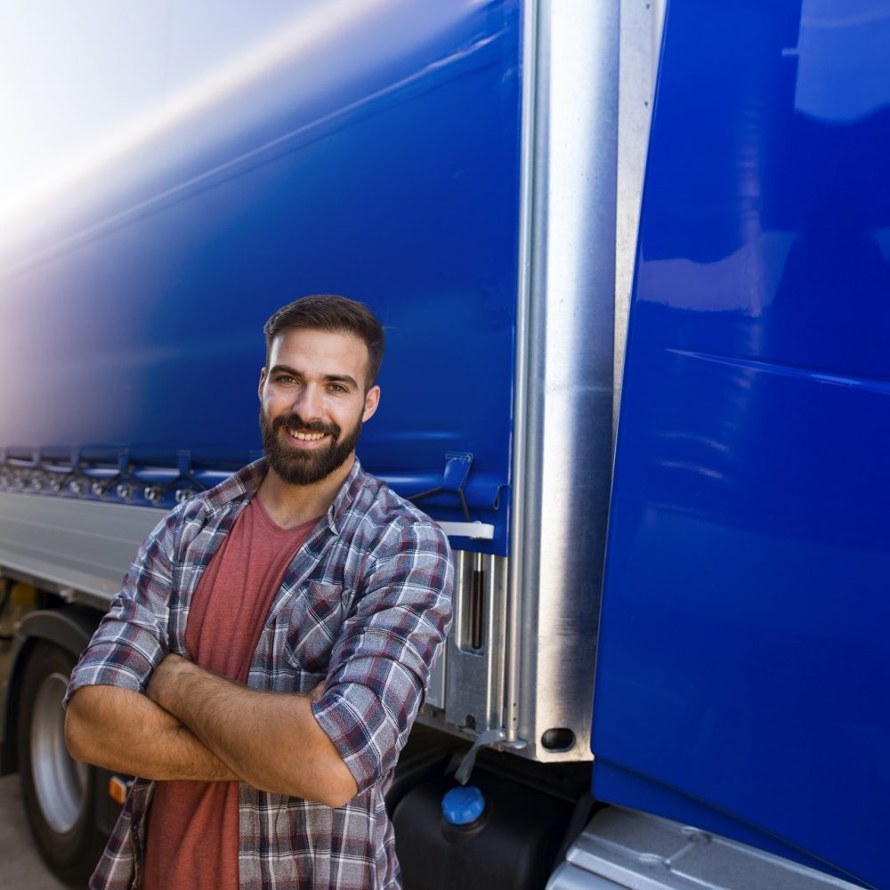 Portrait of professional middle aged bearded truck driver in casual clothes with crossed arms standing by his truck.
